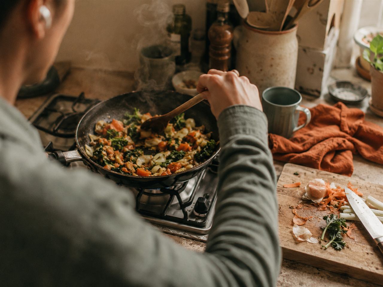 Stirring a pan of vegetables at a home hob with an earbud in, warm evening light